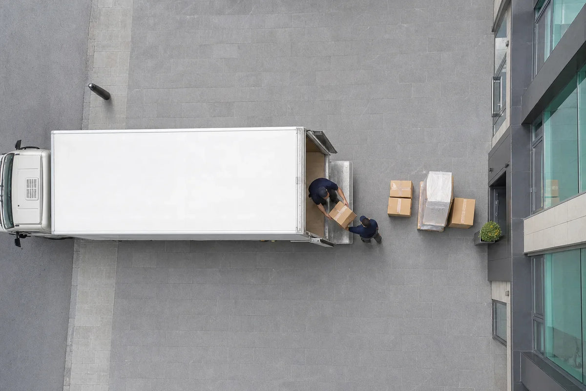 Two movers unloading cardboard boxes from a white delivery truck on a grey tiled driveway beside a glass and metal building