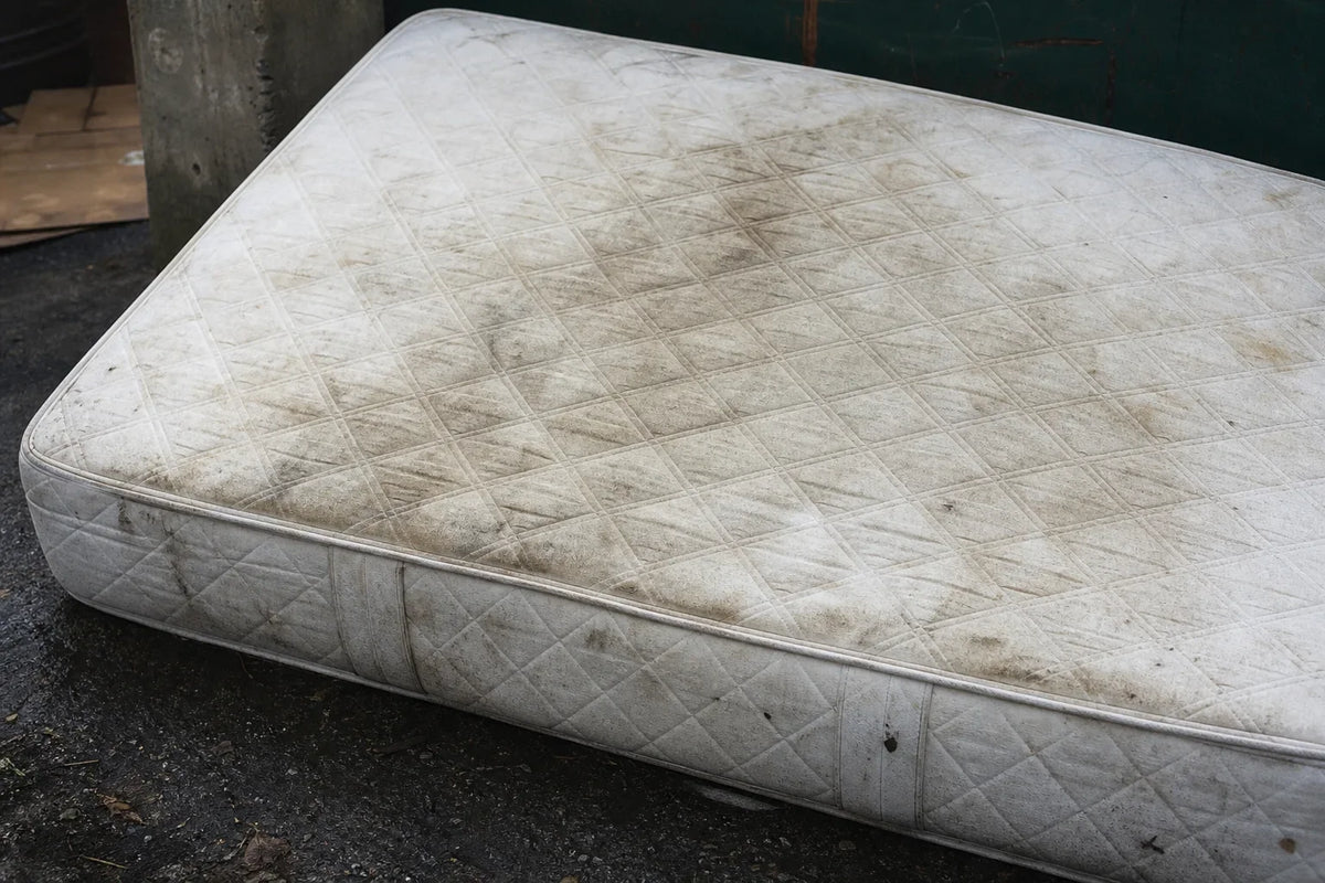 Worn white mattress with dirt marks and stains seen at an angle outdoors on dark pavement